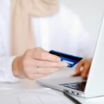 Close-up of a person holding a credit card while using a laptop for online shopping.